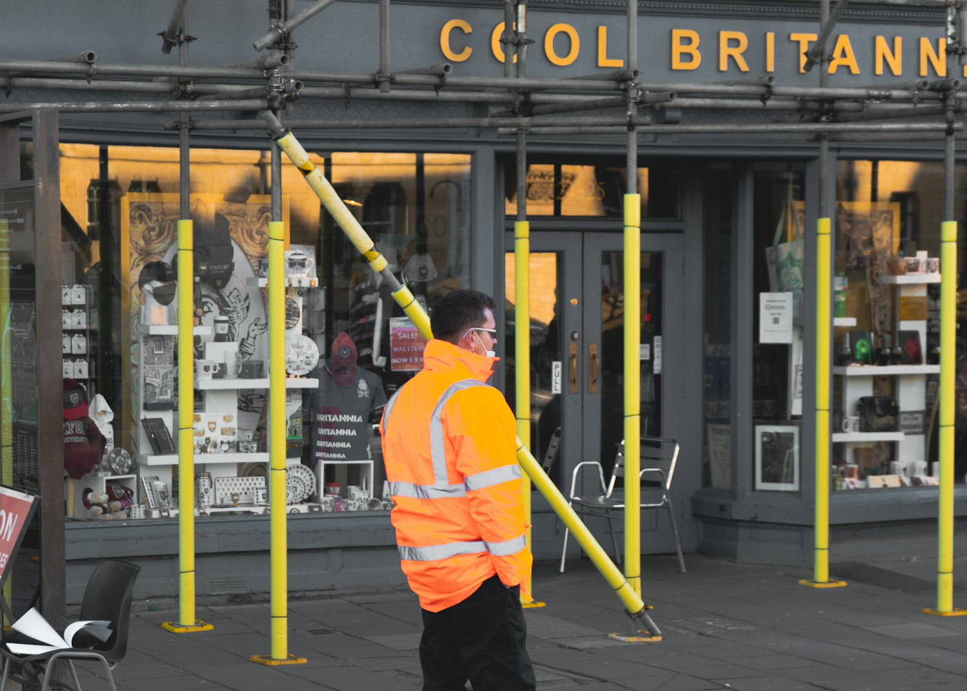 Scaffolder in hi-vis carrying a pole outside a shop on a British high street